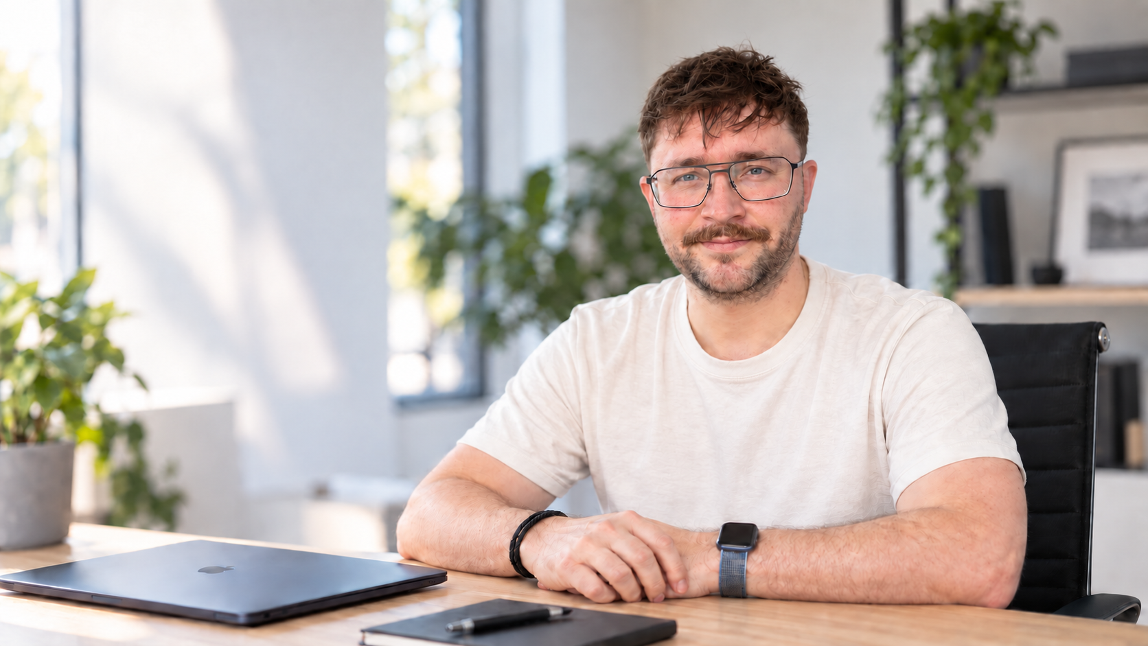 Christoph Hampe sitzt lächelnd an einem Holztisch in einem hellen, modernen Büro. Er trägt ein helles T-Shirt und eine Brille. Vor ihm auf dem Schreibtisch liegen ein geschlossener Laptop und ein Notizblock