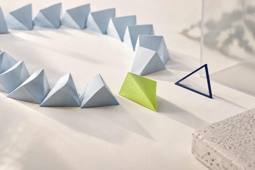 Ein Still-Life-Foto auf hellem Untergrund mit einer Anordnung hellblauer Papier-Pyramiden. Eine einzelne, leuchtend neon-grüne Pyramide steht am Ende der Kette. Rechts davon ein dunkelblauer Dreiecksrahmen. Im Hintergrund Acrylblock- und Blattschatten.