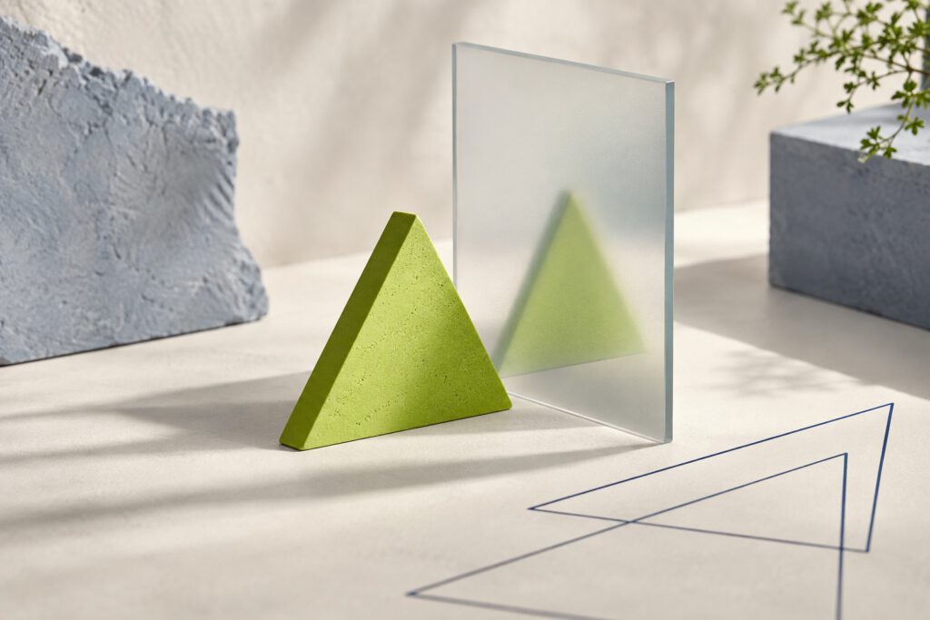 Ein modernes architektonisches Stillleben, das konzeptionelle Marketing-Wirkung und Markenrealität verkörpert. Es zeigt ein zentrales neongrünes Dreieckprisma mit versteinerter Textur, eine große stehende matte Glasplatte mit subtilen Hologrammen von Marketing-Metriken, und ein komplexes 3D-Drahtmodell-Gitter. Betonblöcke und eine Pflanze ergänzen die Szene unter weichem, dappled Licht.